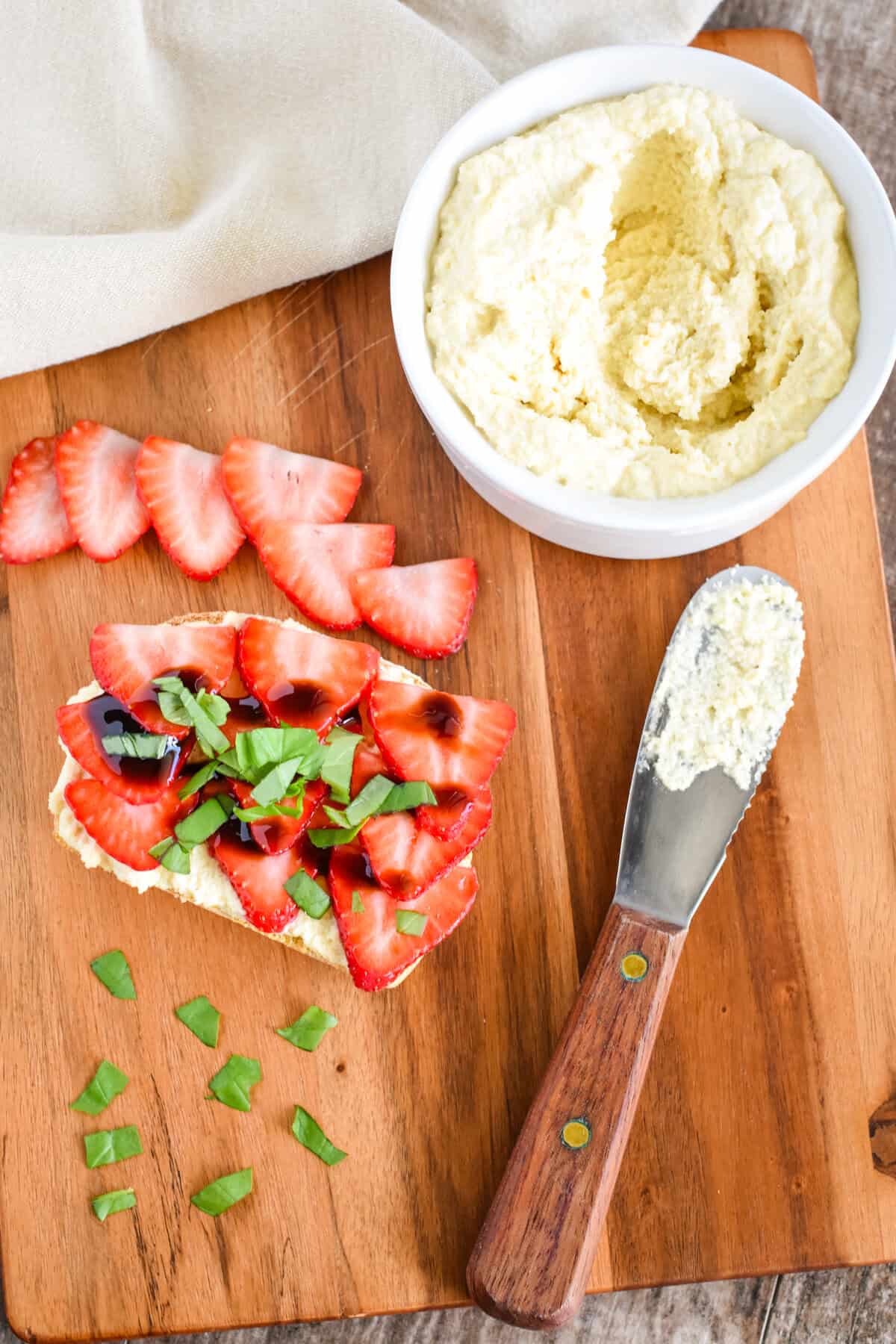 A piece of toast topped with vegan ricotta, sliced strawberries, basil, and balsamic glaze on a wooden board.