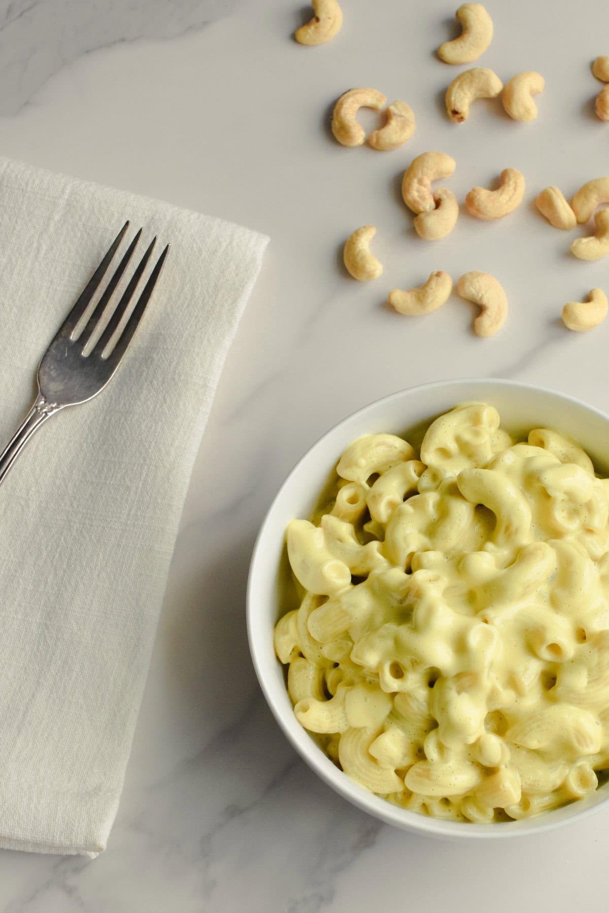 Dairy-free mac and cheese in a white bowl with a fork on a kitchen napkin next to it and cashews behind it.