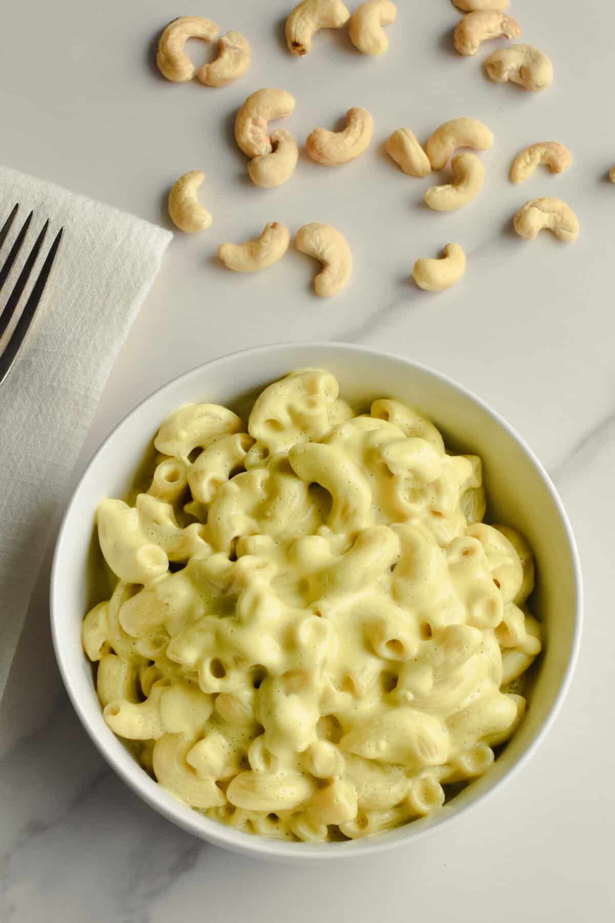 A white bowl filled with vegan mac and cheese on a marble surface with cashews behind it.