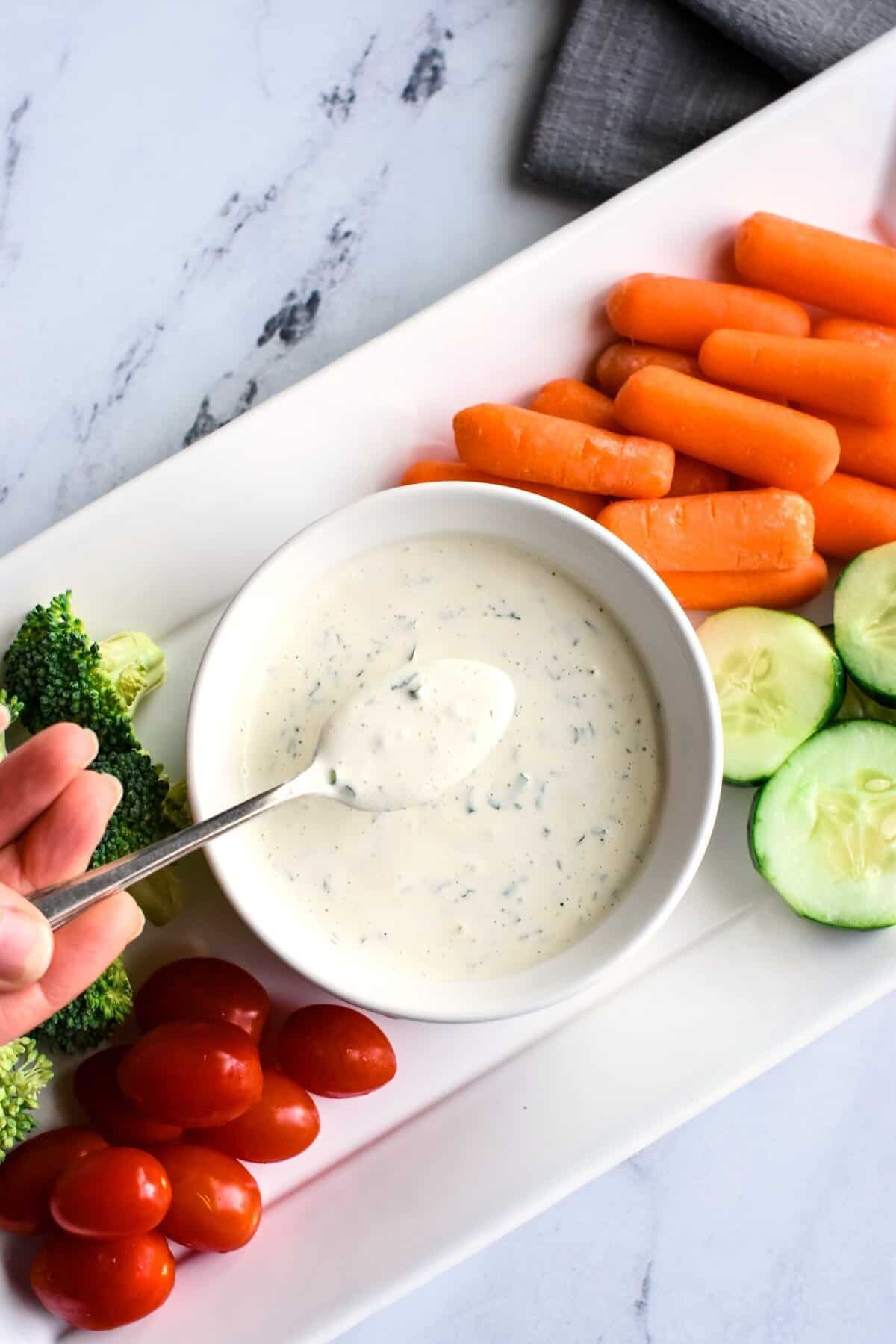 A hand holding a spoonful of dressing over a bowl of it on a platter with veggies.