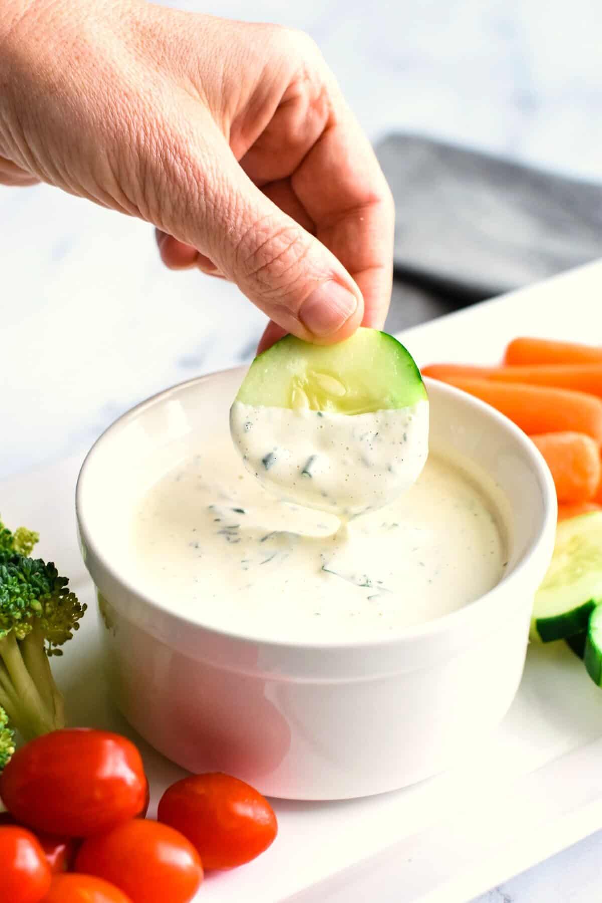 A hand dipping a cucumber slice into a bowl of cashew ranch dressing on a veggie platter.