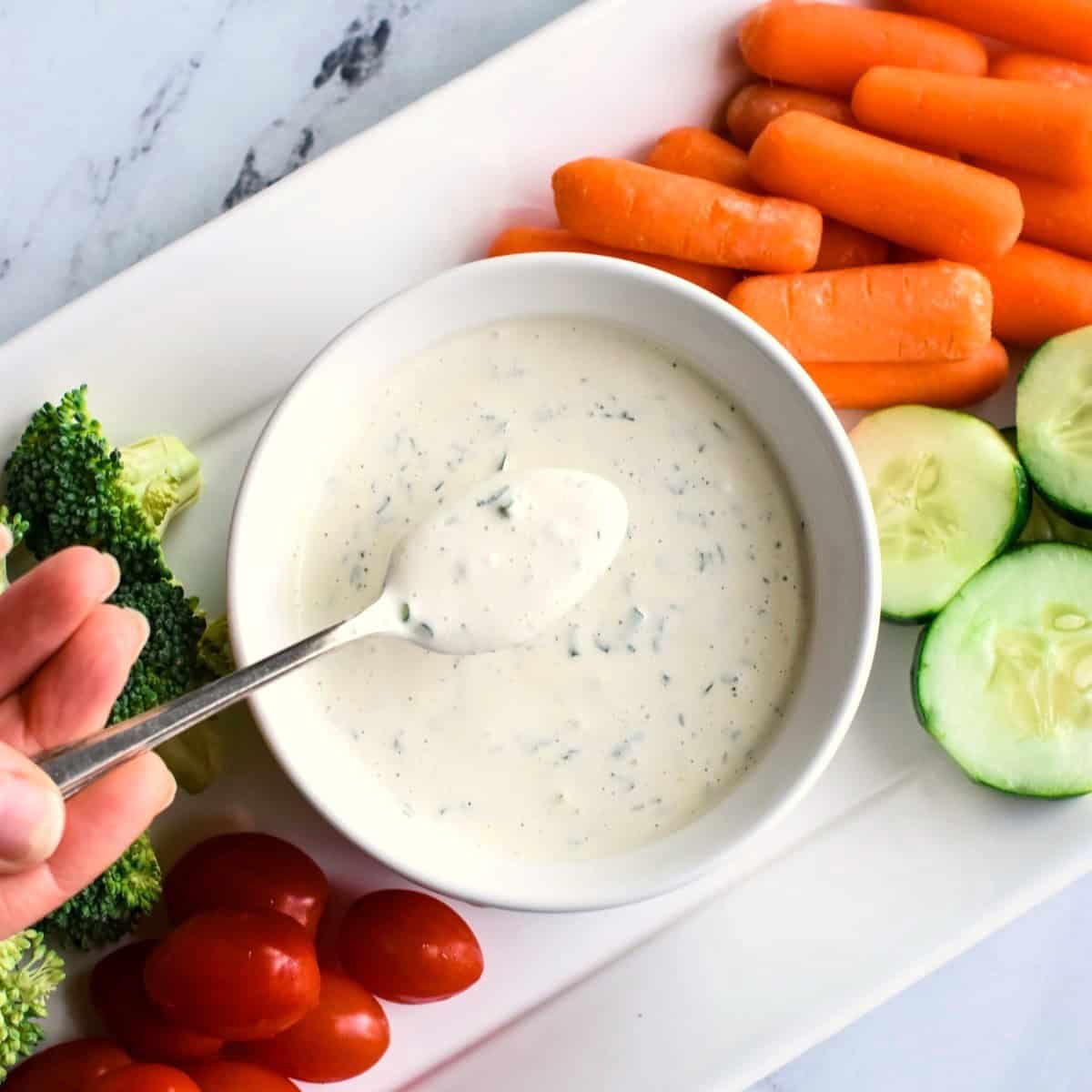 A person holding up a spoon with dressing on it over a bowl with the rest of it on a veggie platter.