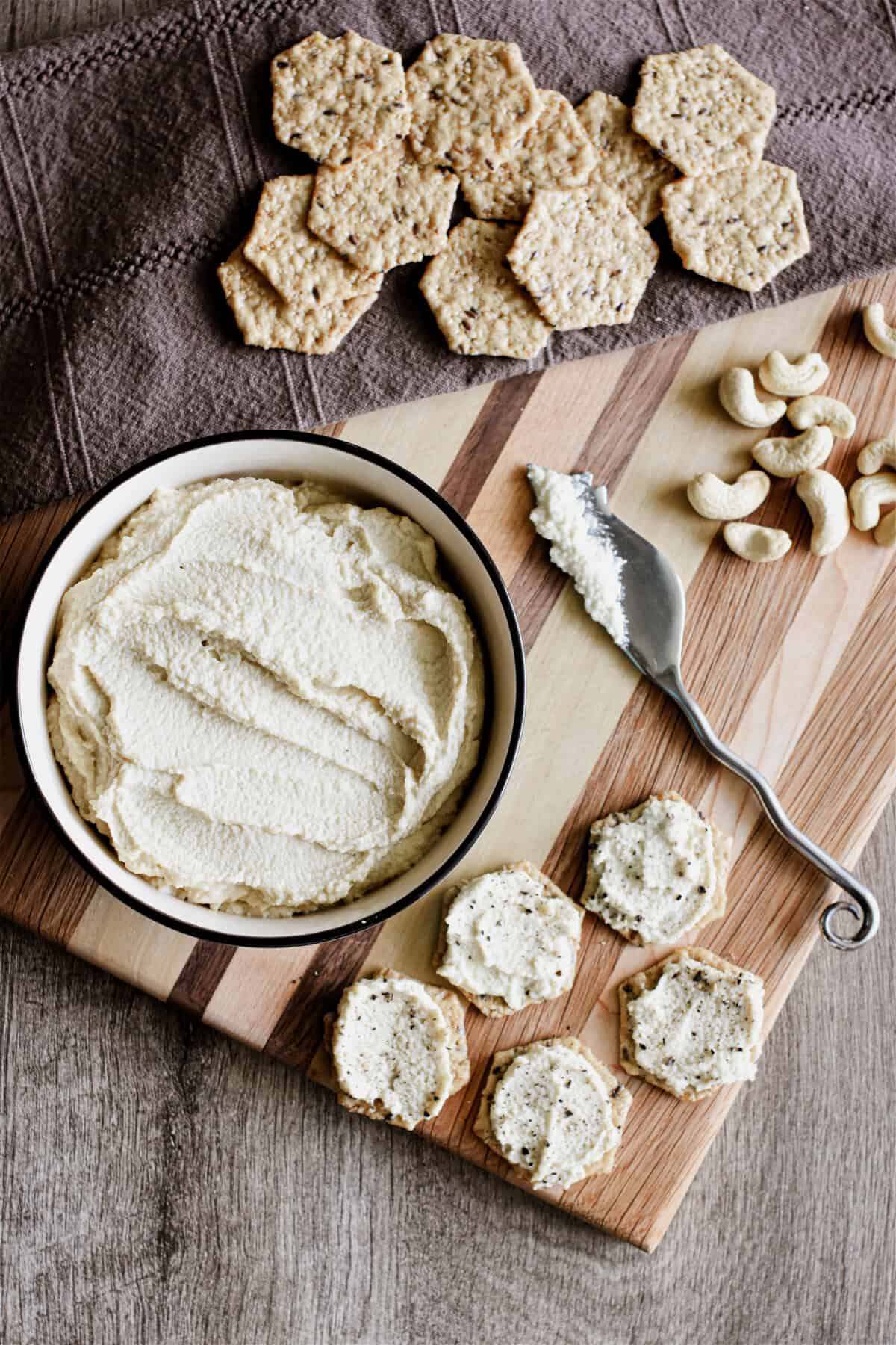 A wooden board with a bowl of cashew ricotta and some crackers topped with the spread.
