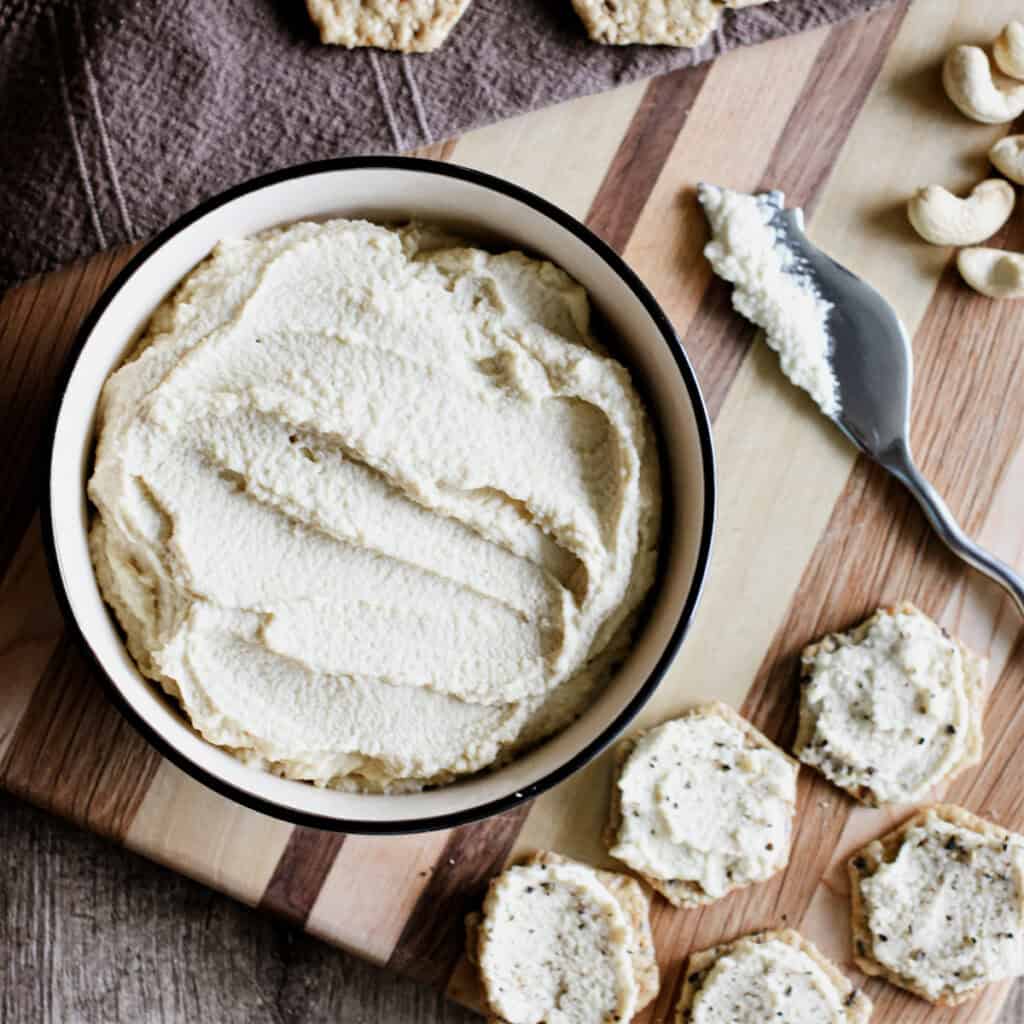 A bowl of cashew ricotta on a wooden board with crackers that have some of it on them.