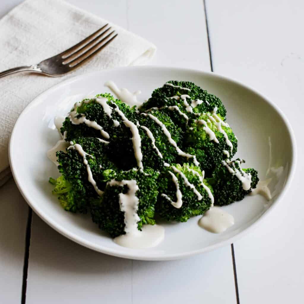A white plate of steamed broccoli drizzled with cashew mozzarella sauce.