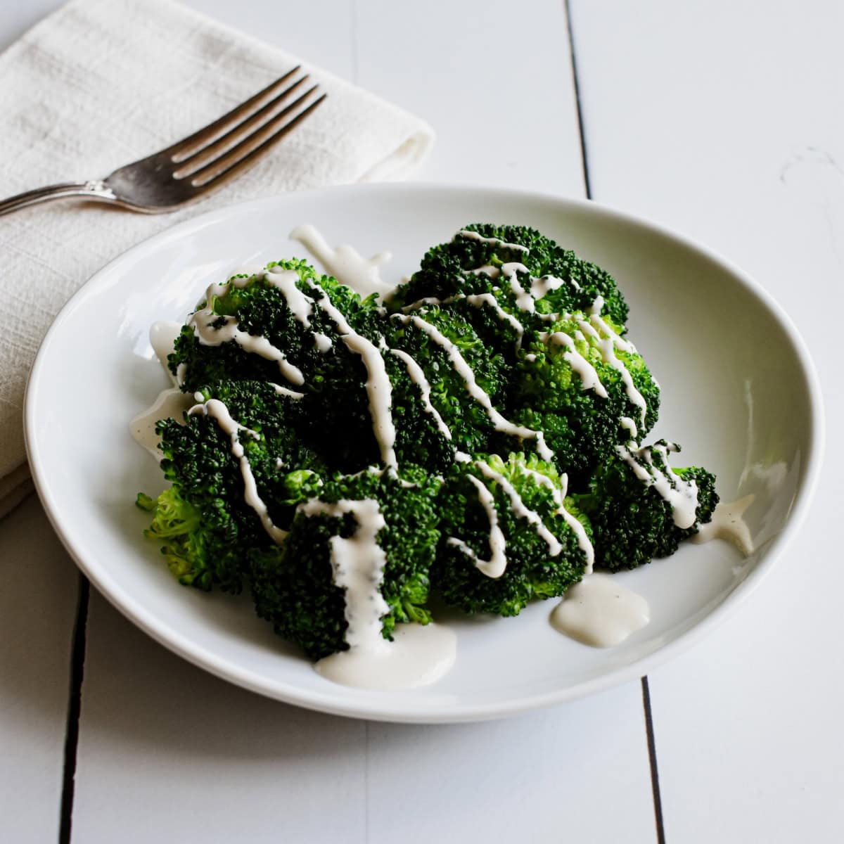 A white plate of steamed broccoli drizzled with cashew mozzarella sauce.