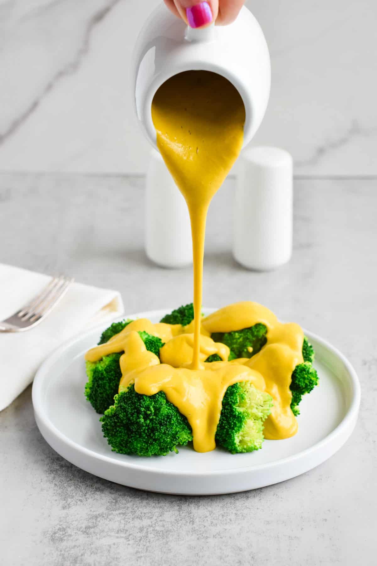 A hand pouring vegan cheese sauce from a container onto a plate of broccoli.
