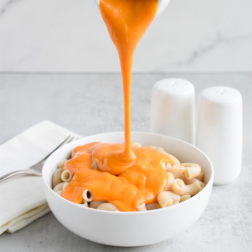 Vegan cheese being poured onto macaroni in a white bowl.