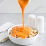 Vegan cheese being poured onto macaroni in a white bowl.